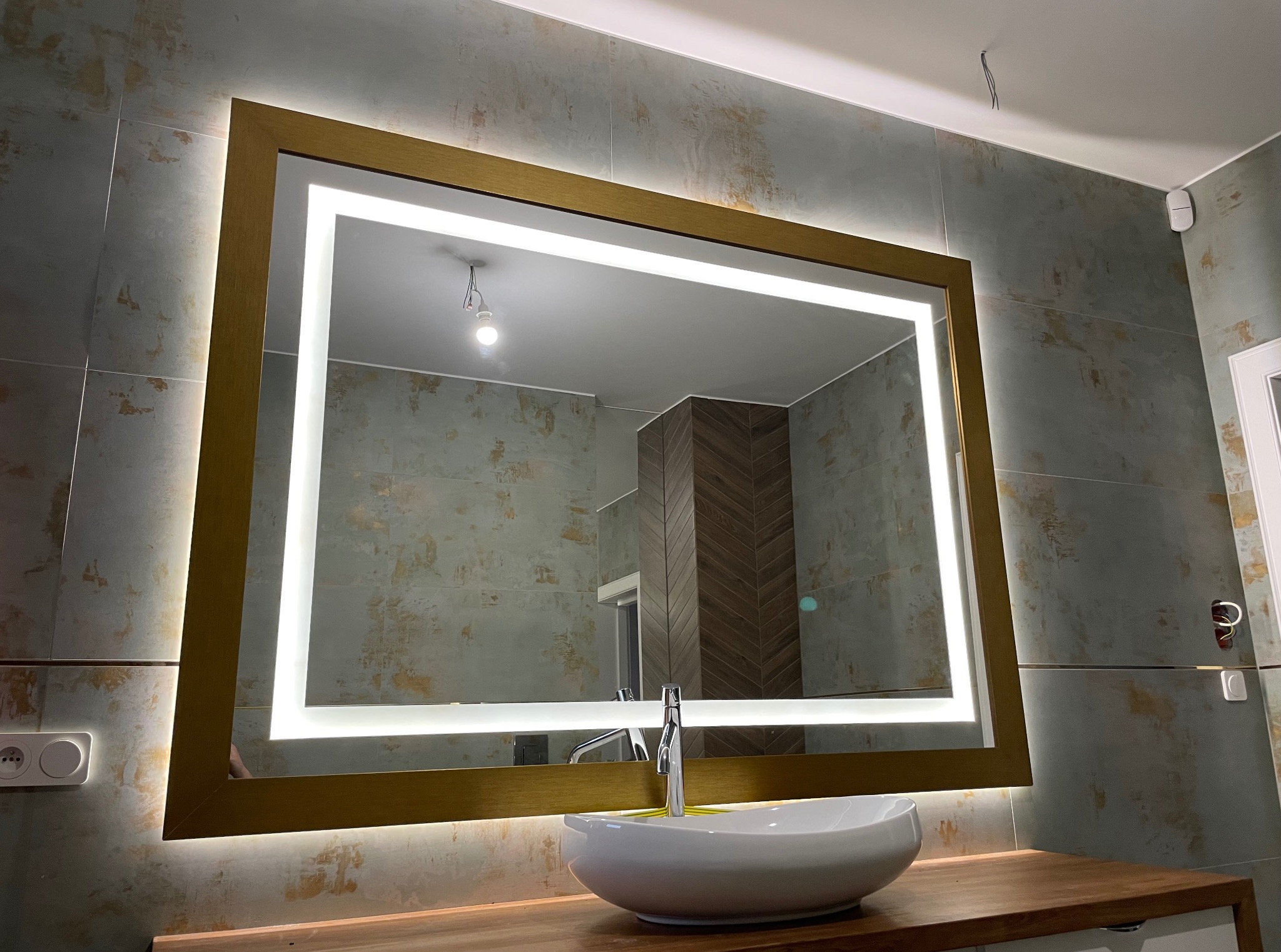 Łazienka z podświetlanym lustrem w złotej ramie, umywalką nablatową i płytkami w odcieniach szarości. Widoczne gniazdko elektryczne i fragment instalacji oświetleniowej.