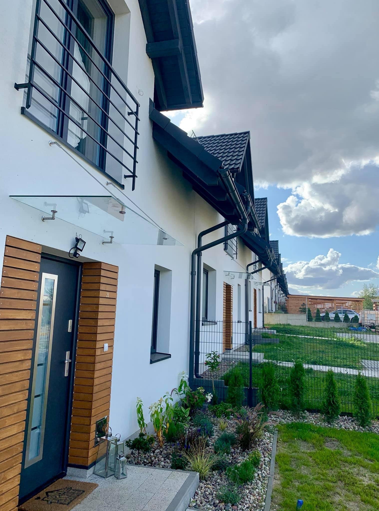 Szereg domów jednorodzinnych z czarnymi balustradami balkonowymi i szklanymi zadaszeniami nad wejściami, w nowoczesnym stylu, z elementami drewnianymi.