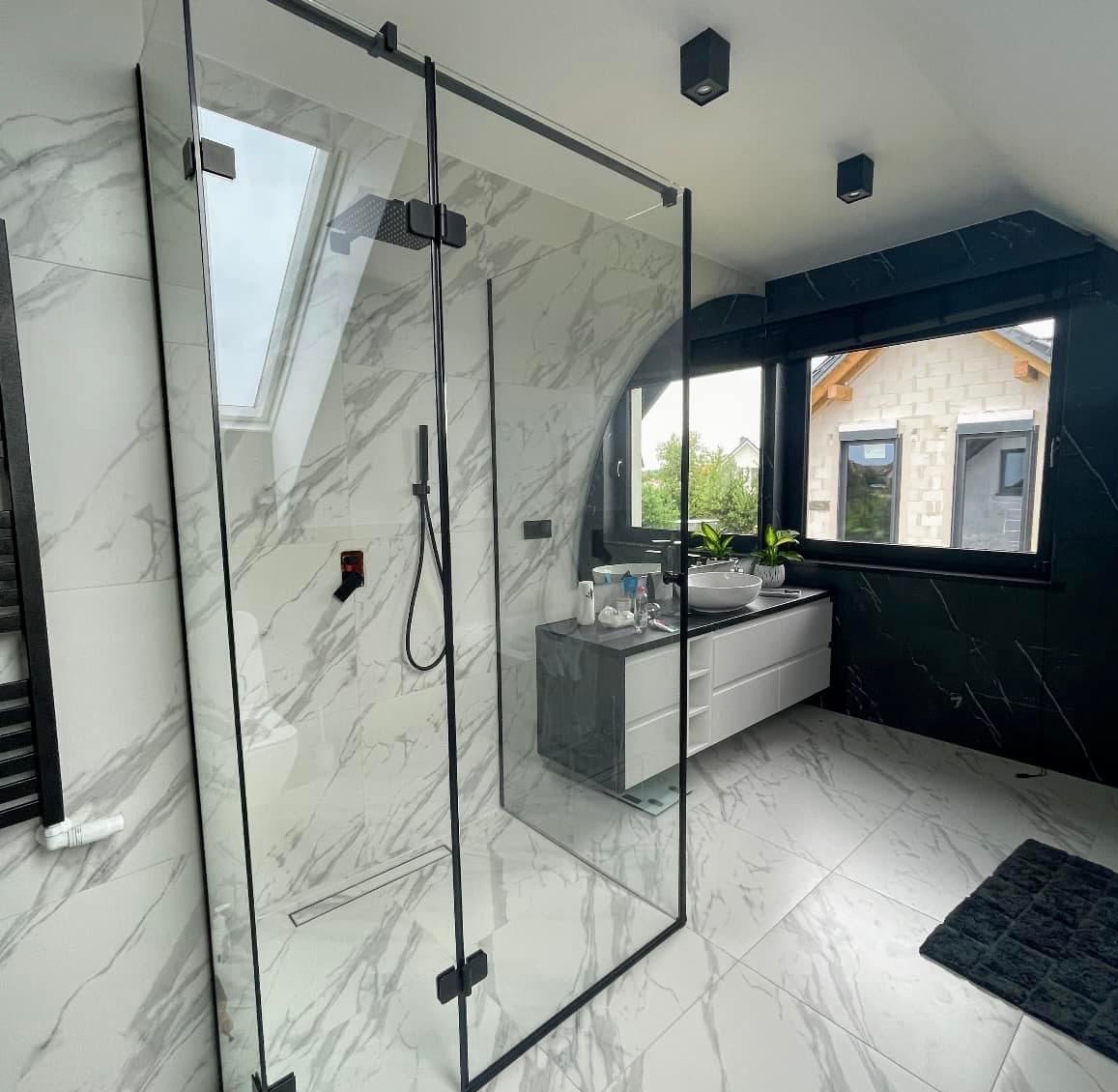 Nowoczesna kabina prysznicowa z czarnymi profilami na tle marmurowej łazienki z oknem i skośnym sufitem. Widok na umywalkę i budynek w budowie za oknem.