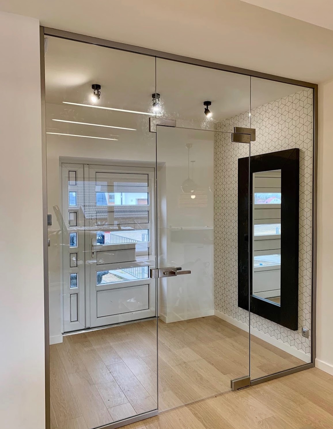 Szklana ściana działowa z drzwiami, metalowa rama, widok na korytarz i drzwi wejściowe. Nowoczesny design, naturalne światło, drewniana podłoga, geometryczna tapeta.