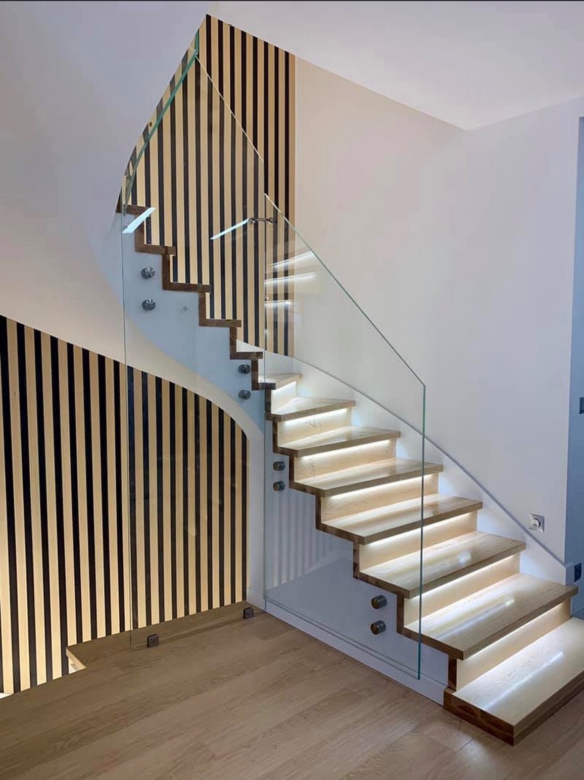 Nowoczesne schody drewniane z podświetleniem LED i szklaną balustradą, zamocowaną na metalowych uchwytach. Ściana z dekoracyjnymi panelami z pionowymi listwami.