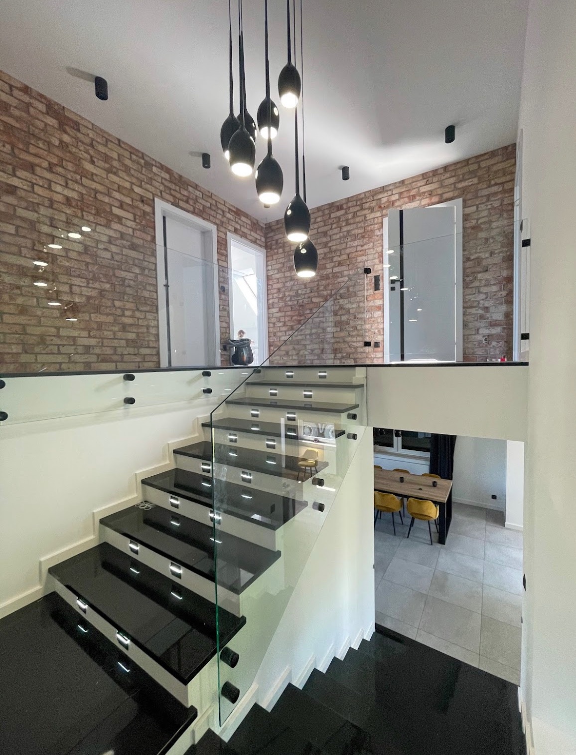 Nowoczesne schody z czarnymi stopniami i szklaną balustradą, oświetlone wiszącymi lampami, na tle ceglanej ściany i widoku na jadalnię. Eleganckie wykończenie wnętrza.