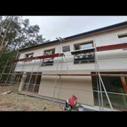 Elewacja domu po tynkowaniu w Grodzisku Mazowieckim. Widoczne rusztowania i osoba pracująca przy elewacji. Część ściany bez tynku, reszta w jasnym odcieniu.