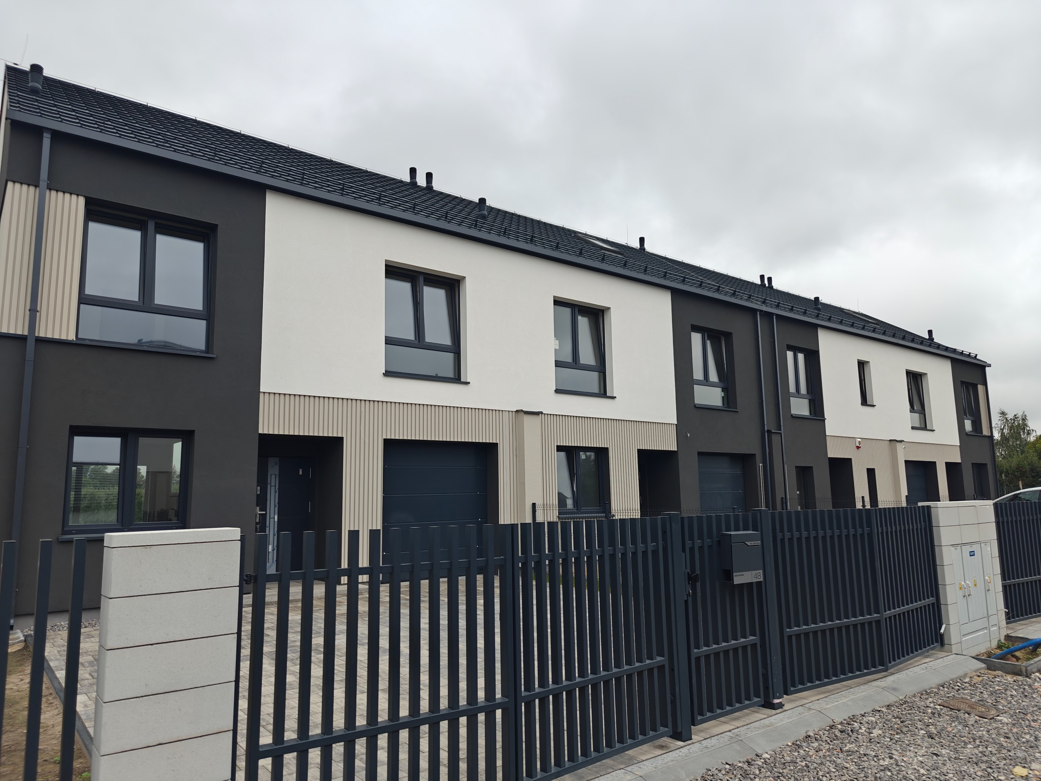 Szereg domów jednorodzinnych z płaskim dachem, w nowoczesnej kolorystyce: szaro-białe elewacje, grafitowa stolarka i ogrodzenie panelowe. Widok z zewnątrz.