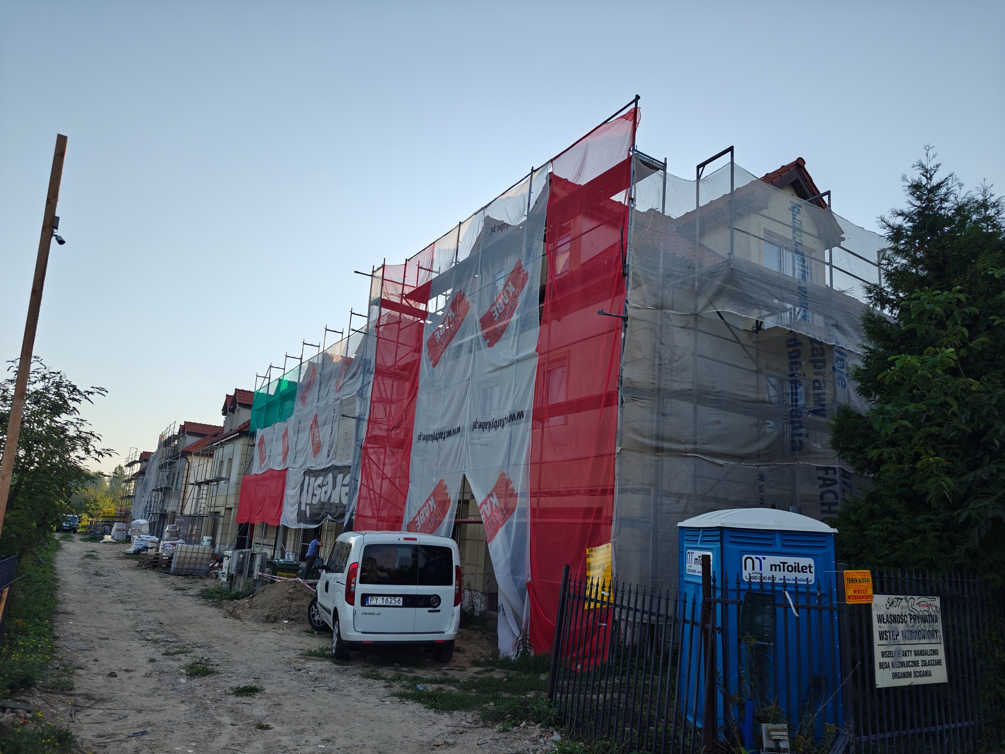 Budynek w rusztowaniach osłonięty siatką ochronną z reklamami, widoczny z drogi gruntowej. Na pierwszym planie toaleta przenośna i zaparkowany biały samochód dostawczy.