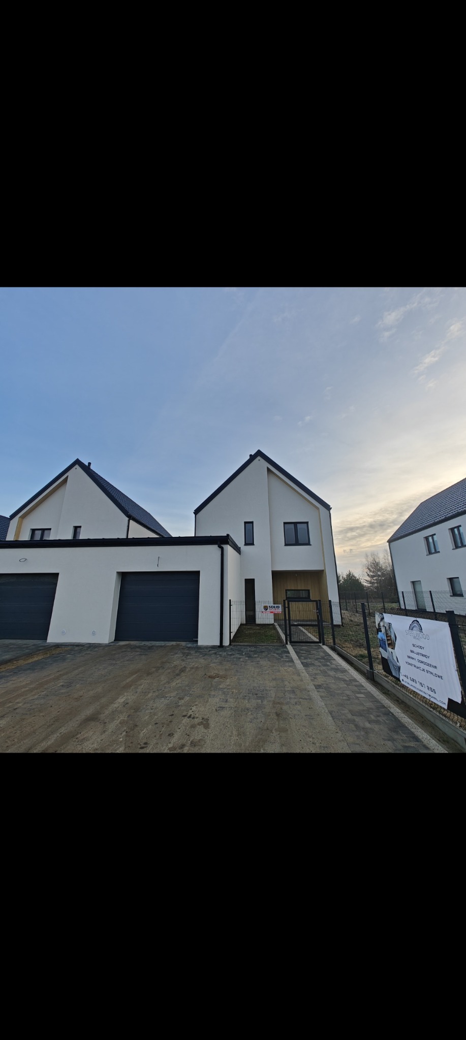 Nowoczesne, białe domy jednorodzinne z grafitowymi akcentami (dachy, okna, bramy garażowe) w słoneczny dzień. Widoczny chodnik z kostki i ogrodzenie.