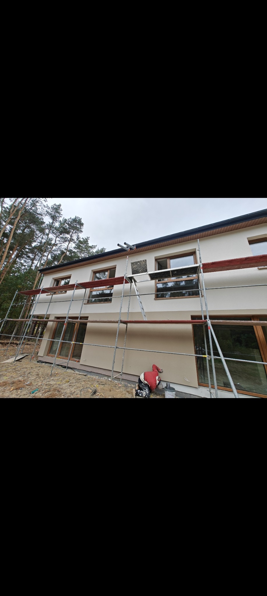 Elewacja domu po tynkowaniu w Grodzisku Mazowieckim. Widoczne rusztowania i osoba pracująca przy elewacji. Część ściany bez tynku, reszta w jasnym odcieniu.