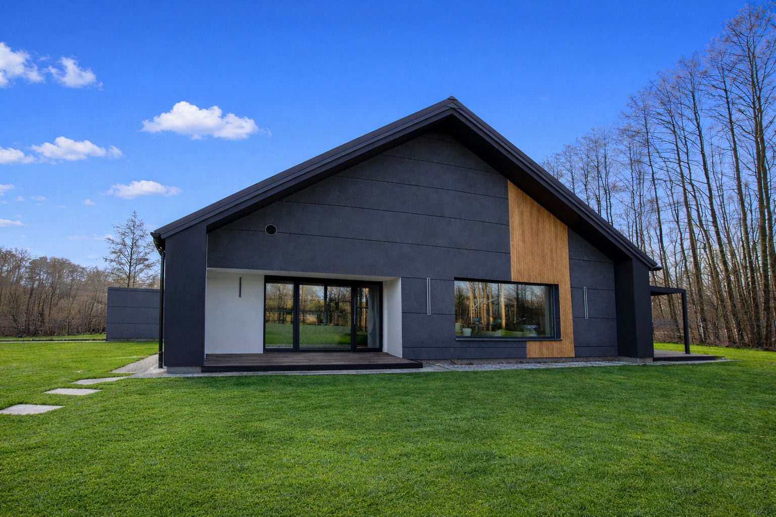 Nowoczesny dom jednorodzinny z grafitową elewacją, drewnianymi akcentami i dużymi oknami, otoczony zielonym trawnikiem i drzewami. Błękitne niebo z chmurami w tle.