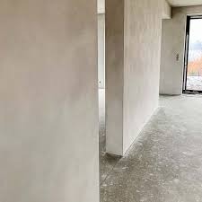 Gładkie, jasnoszare tynki maszynowe w nowym domu w Niechlowie. Widok na surową podłogę i otwarte drzwi balkonowe. Minimalistyczne wykończenie ścian.