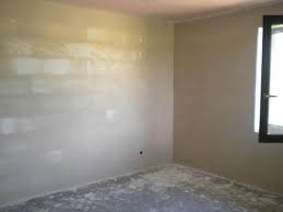 Wnętrze pokoju po tynkowaniu maszynowym, widoczne gładkie, jasne ściany, okno z ciemną ramą i resztki materiału na podłodze. Delikatne światło dzienne.