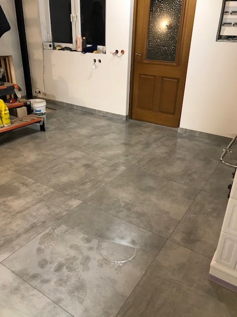 Ułożone szare płytki podłogowe w pomieszczeniu z oknem i drewnianymi drzwiami. Widoczne ślady po pracach wykończeniowych. Na ścianie gniazdka elektryczne.