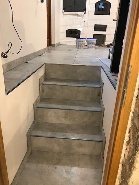 Betonowe schody z metalowymi listwami wykończeniowymi, prowadzące do pomieszczenia z piecem kaflowym. Widok zza framugi drzwi.
