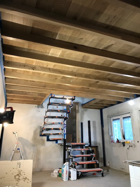 Wnętrze w trakcie wykańczania: drewniany sufit z belek, spiralne schody w metalowej konstrukcji, okno i ściany przygotowane do malowania. Etap prac wykończeniowych.