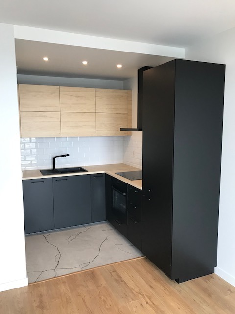 Nowoczesna, kompaktowa kuchnia z szarymi szafkami dolnymi, drewnopodobnymi górnymi i czarnymi akcentami. Płytki imitujące marmur na podłodze. Oświetlenie punktowe.