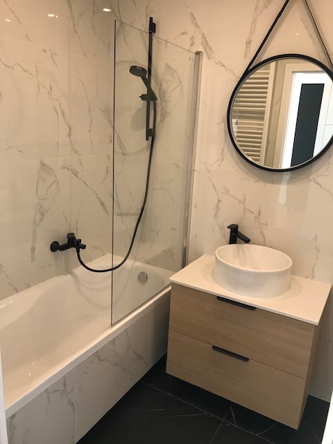 Nowoczesna łazienka z wanną i kabiną prysznicową, białą umywalką nablatową na szafce z jasnego drewna i czarną armaturą. Marmurowe płytki na ścianach.