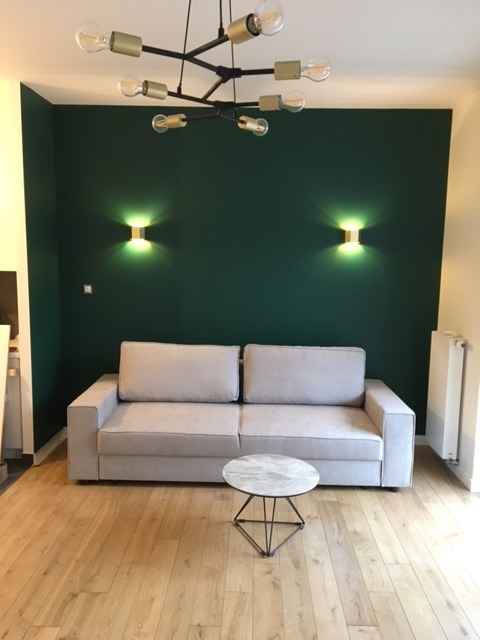 Salon z szarą sofą i stolikiem na tle ciemnozielonej ściany z oświetleniem punktowym. Parkiet z jasnego drewna i nowoczesna lampa sufitowa.