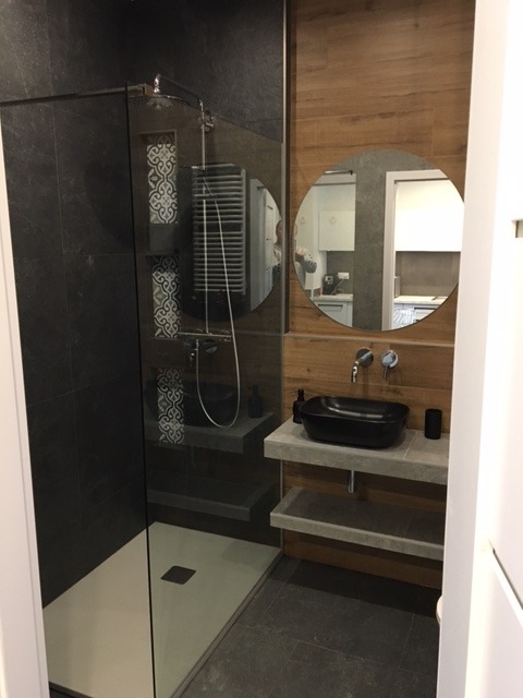 Nowoczesna łazienka z kabiną prysznicową typu walk-in, umywalką nablatową i okrągłym lustrem, wykończona w odcieniach szarości, bieli i drewna.