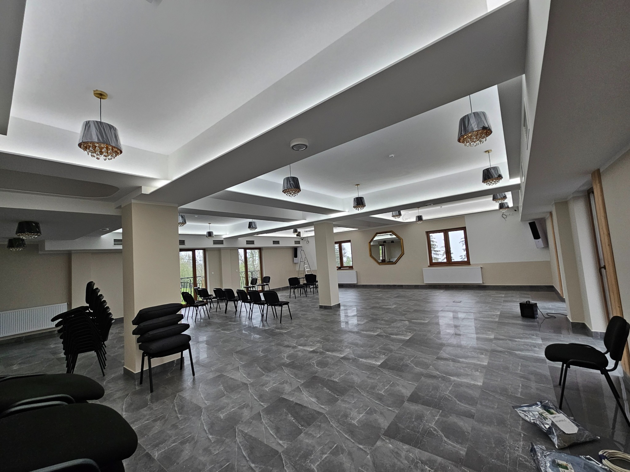 Duża sala konferencyjna z szarymi, kryształowymi lampami, marmurową podłogą i czarnymi krzesłami. Widoczne okna i lustro na ścianie. Oświetlenie sufitowe.