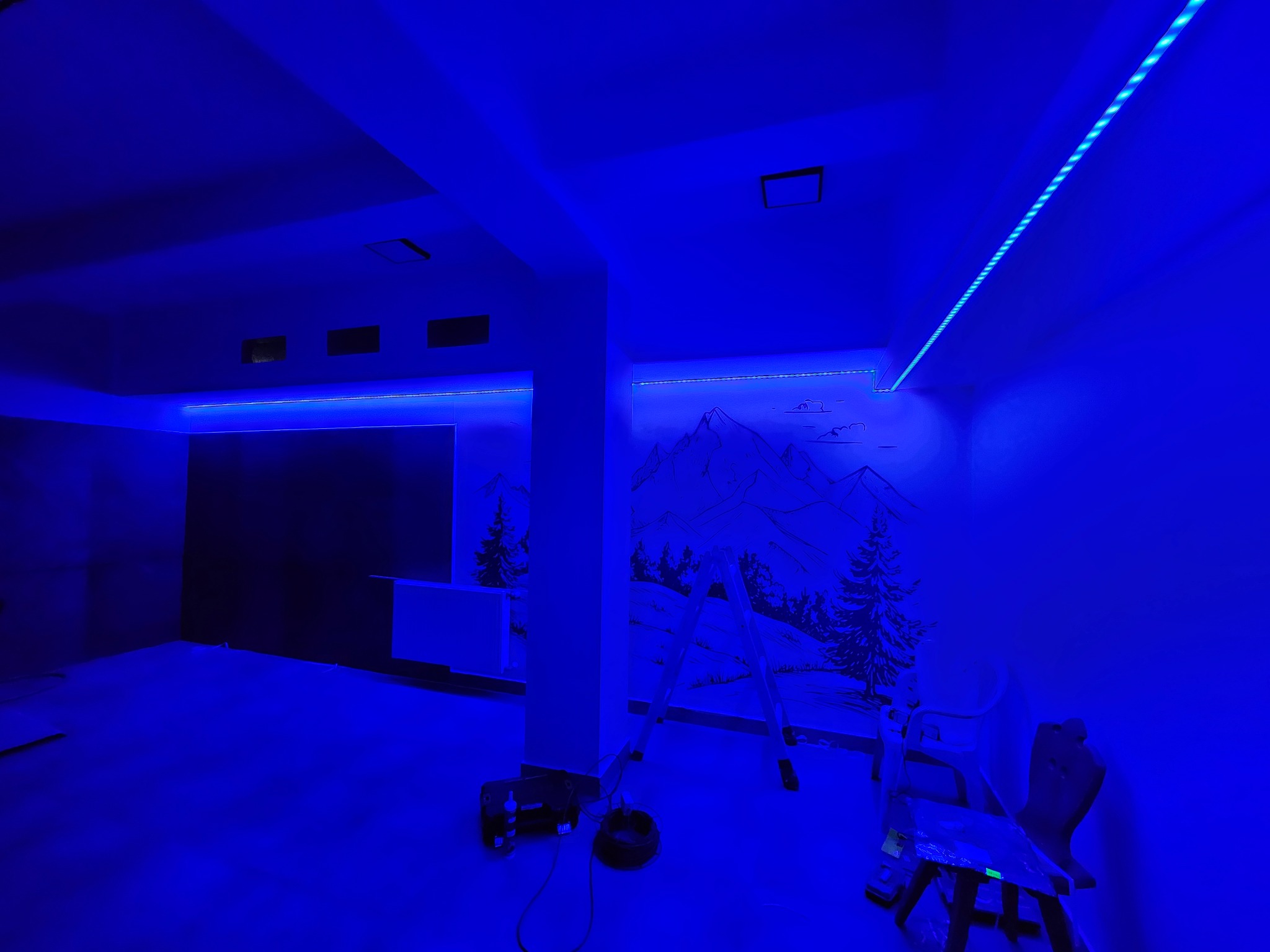 Pokój oświetlony niebieskimi taśmami LED, z rysunkiem gór na ścianie. Widoczne drabina, krzesło i narzędzia, co sugeruje montaż oświetlenia w trakcie realizacji.