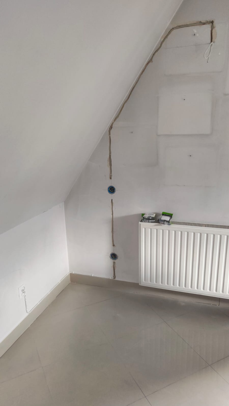 Poddasze z instalacją elektryczną: kable prowadzone po ścianie, gniazdka w puszkach. Widoczny grzejnik i surowy stan wykończenia wnętrza.