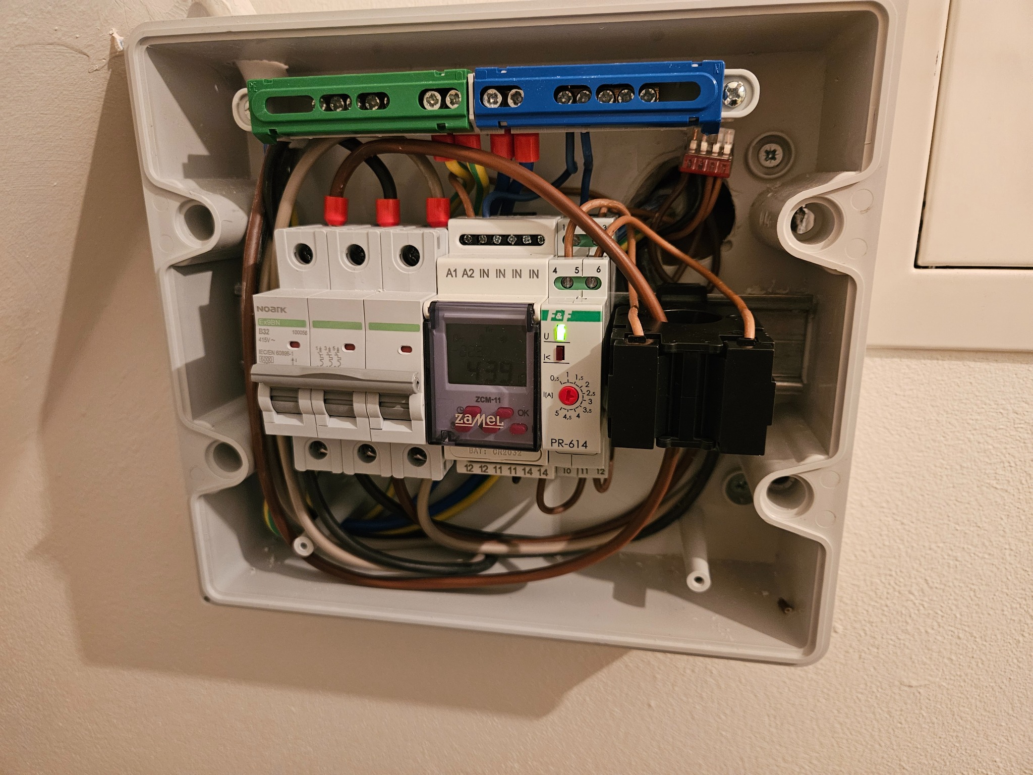 Otwarta skrzynka elektryczna z bezpiecznikami, przewodami i licznikiem Zamel, zamontowana na ścianie. Widoczne listwy zaciskowe w kolorach zielonym i niebieskim.