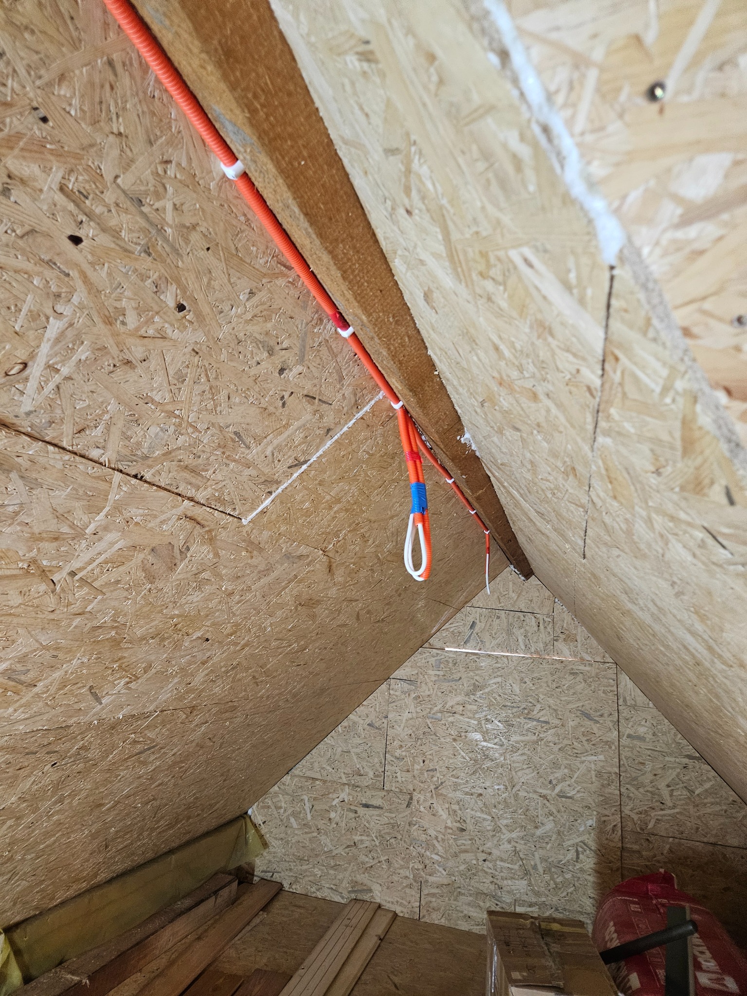 Pomarańczowy przewód elektryczny przymocowany do drewnianej konstrukcji dachu na poddaszu, zabezpieczony białymi uchwytami. Widoczne płyty OSB.