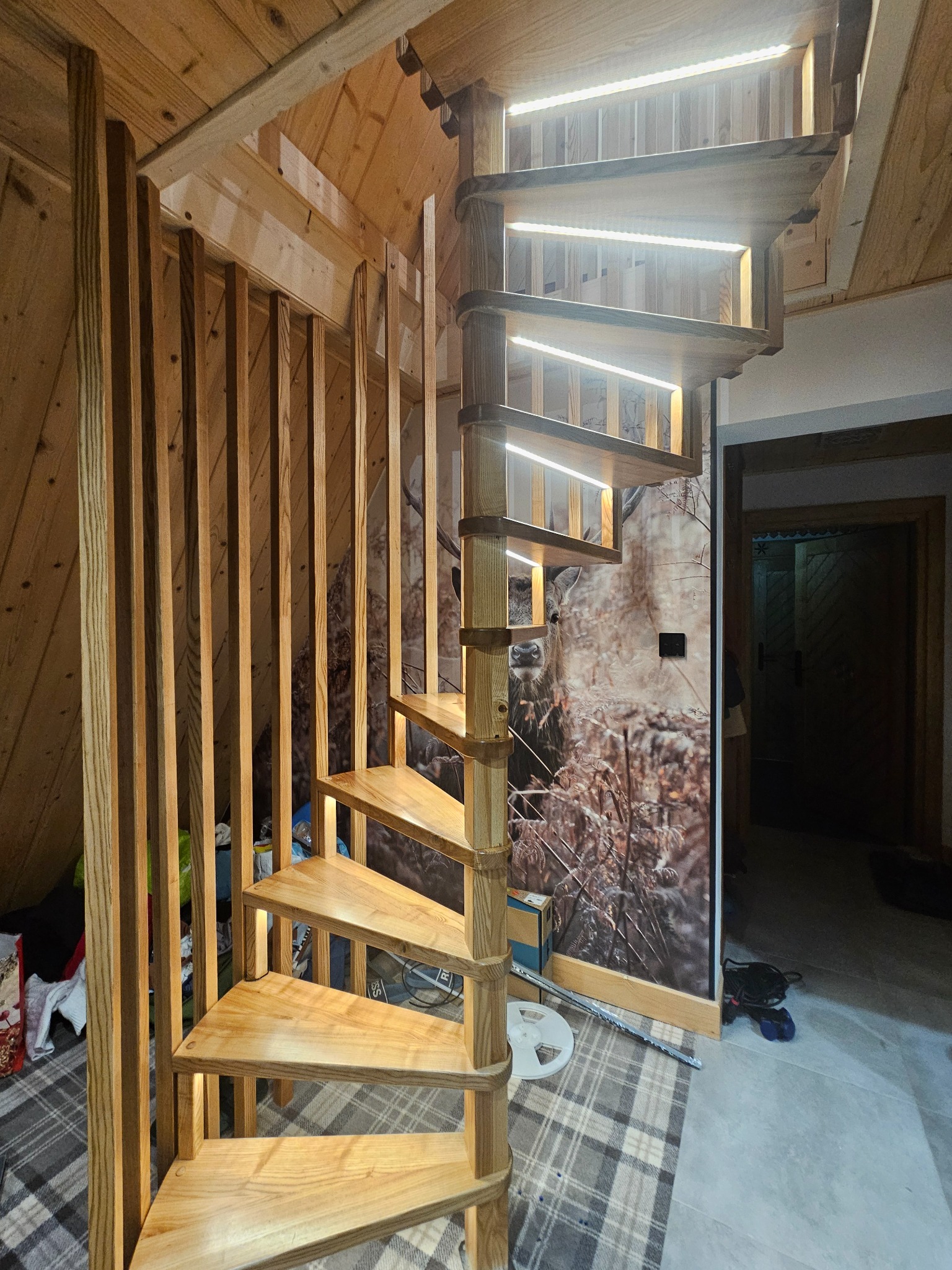 Drewniane schody spiralne z oświetleniem LED, widok z dołu. Naturalne drewno, ciepłe światło, dekoracyjna ściana z motywem leśnym. Element wystroju wnętrza.