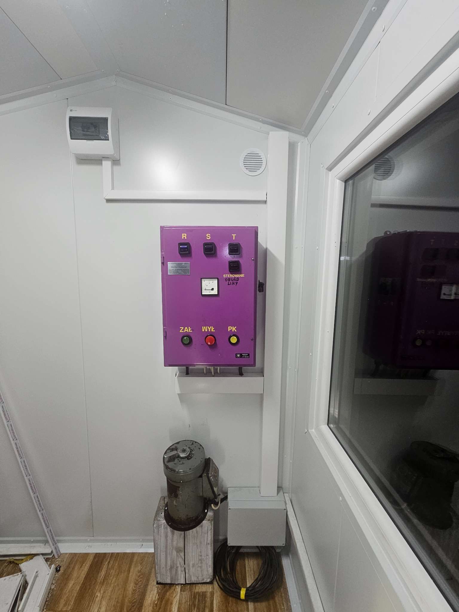 Fioletowa szafa sterownicza z przyciskami R, S, T, ZAL, WYŁ, PK w pomieszczeniu z oknem i elementami instalacji elektrycznej. Widoczne kable i skrzynka przyłączeniowa.