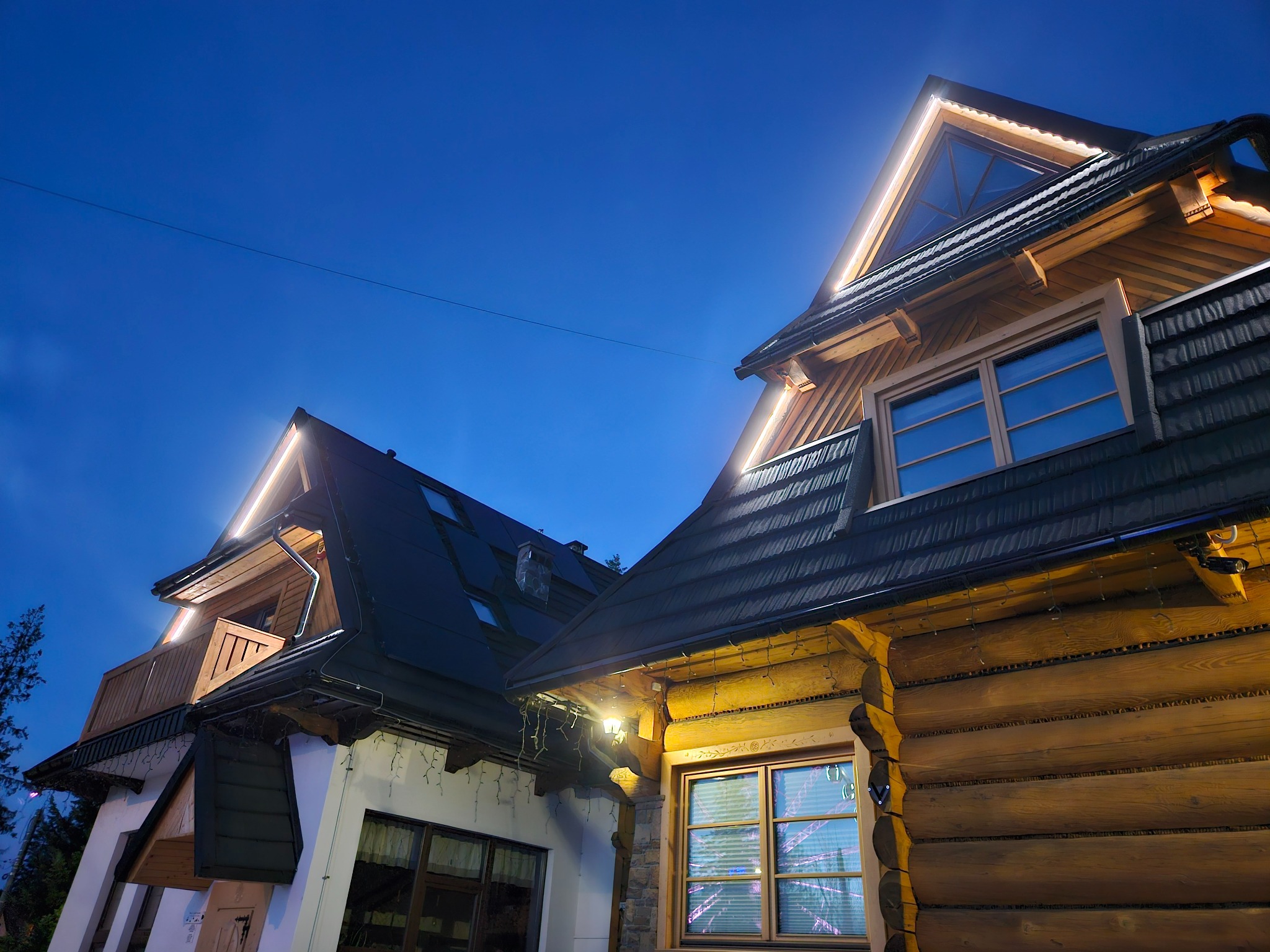 Oświetlony nocą dom w stylu góralskim z drewnianymi elementami i oknami. Na dachu widoczne lampki, tworzące ciepły, klimatyczny efekt.