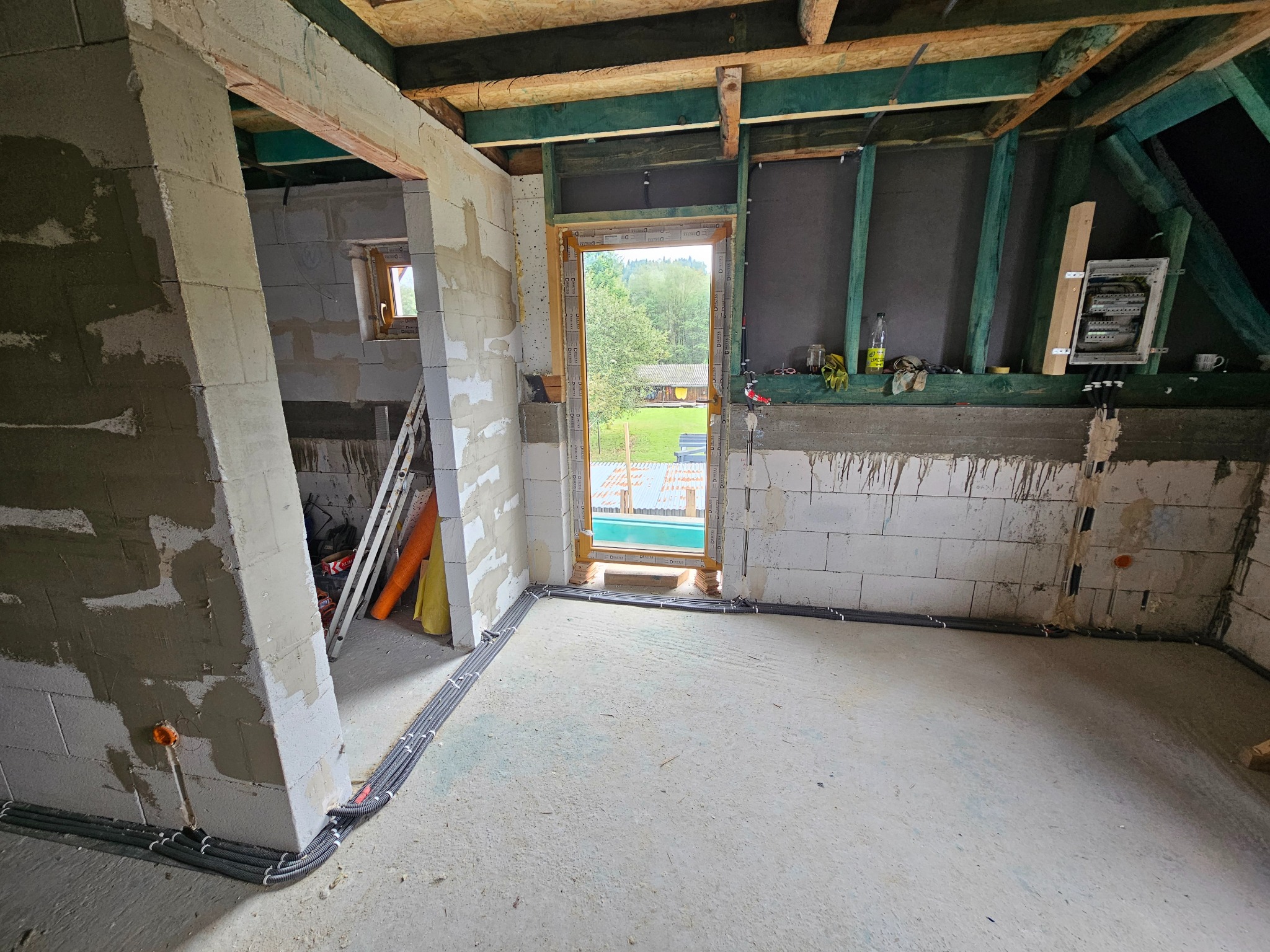 Wnętrze w trakcie budowy z widocznymi przewodami elektrycznymi ułożonymi wzdłuż ścian i podłogi. Na ścianie zamontowana skrzynka elektryczna. Widok na zewnątrz przez otwór okienny.