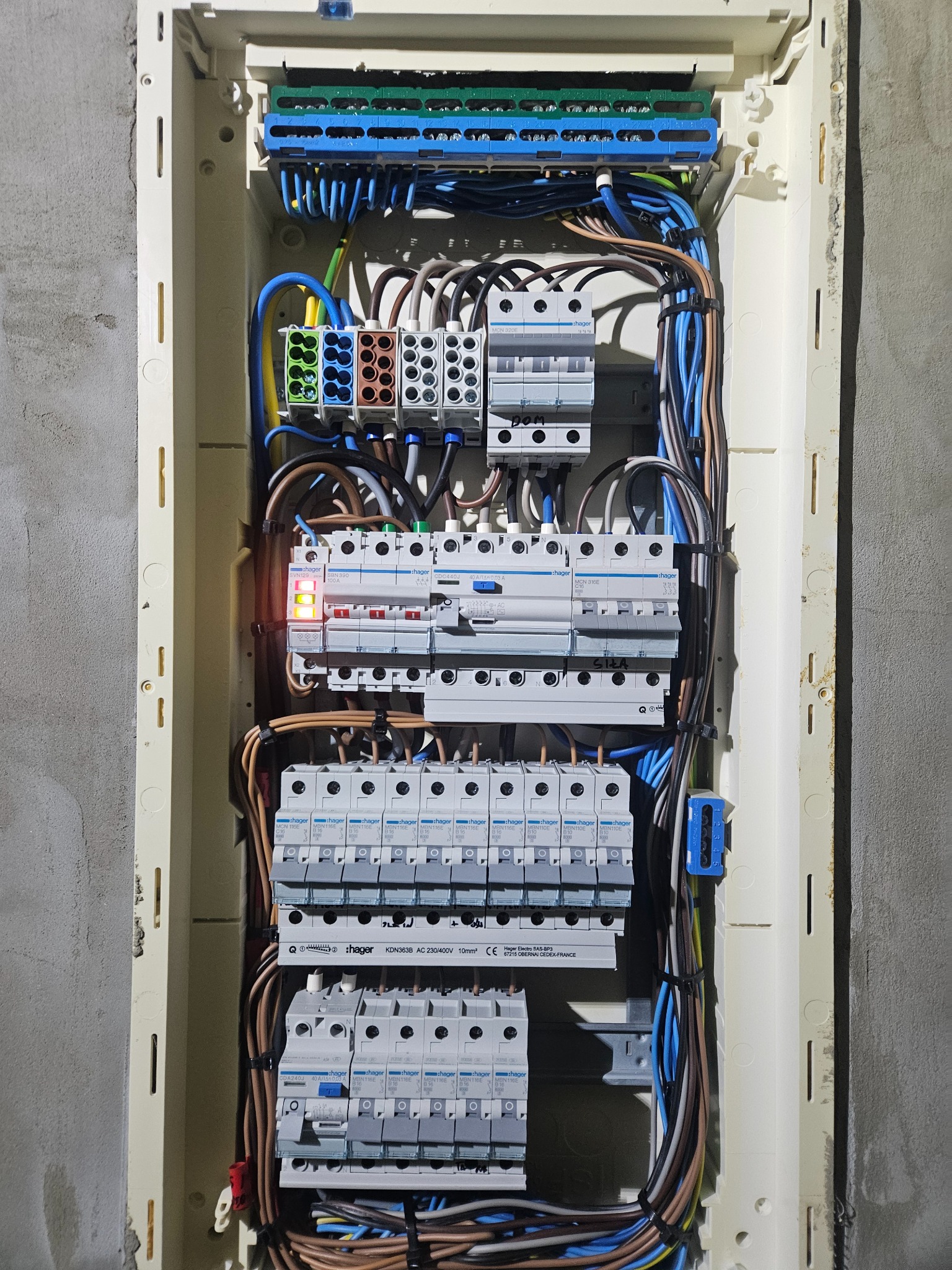 Rozdzielnica elektryczna Hager z zainstalowanymi bezpiecznikami i okablowaniem, widoczna na tle surowej ściany. Jeden z bezpieczników sygnalizuje aktywność.