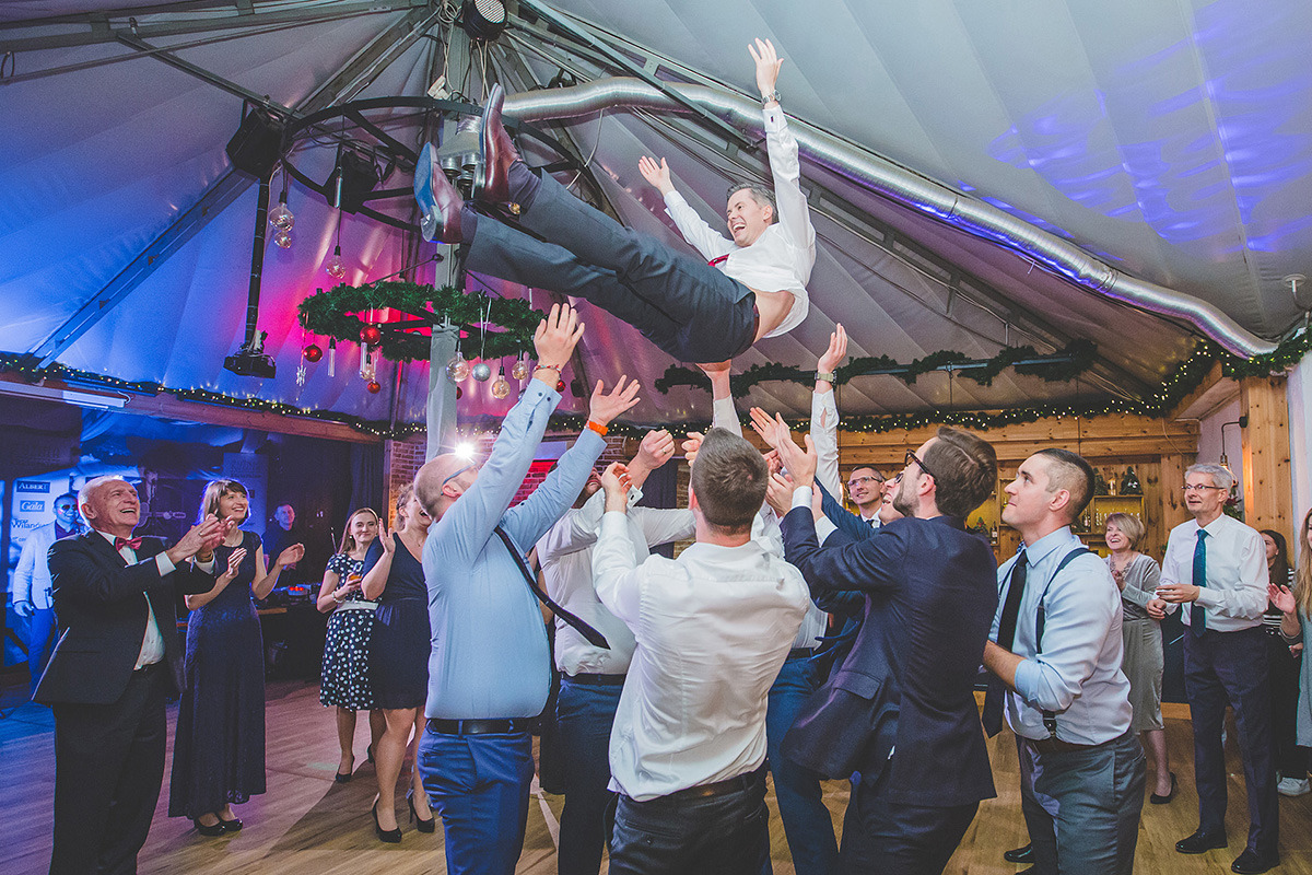 Radosna scena weselna: Pan młody uniesiony w górę przez gości, w tle uśmiechnięci ludzie i dekoracje świąteczne, tworząc dynamiczne i pełne emocji ujęcie.