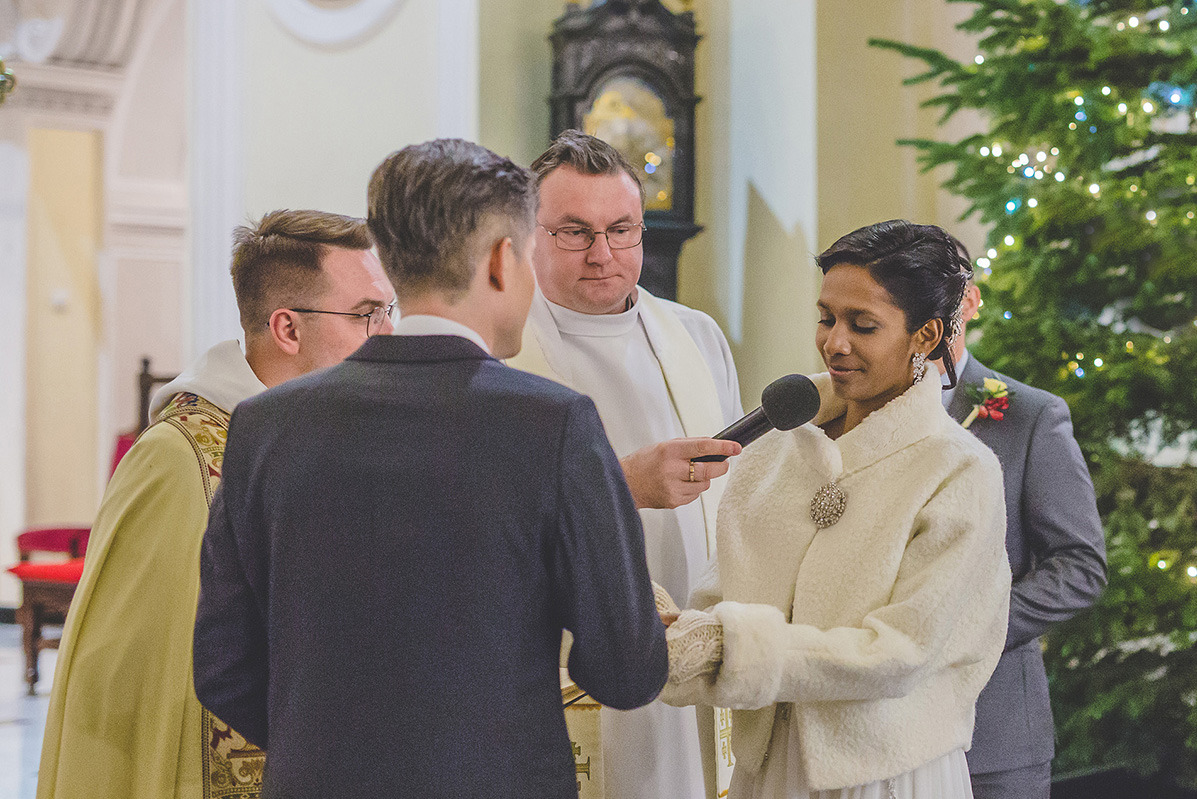 Ślub w kościele: panna młoda z mikrofonem, ksiądz udzielający ślubu, goście w tle, dekoracja choinkowa. Ujęcie z bliska, ciepłe oświetlenie.
