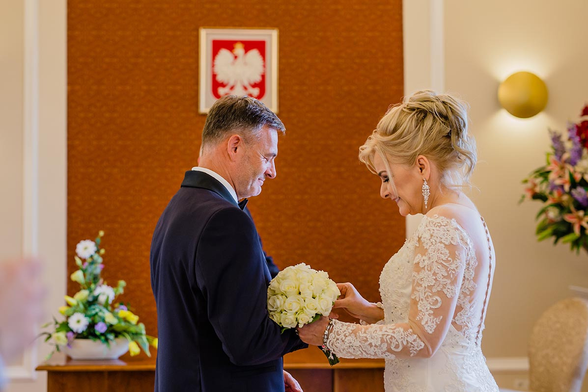 Para młoda wymienia się obrączkami w urzędzie stanu cywilnego. W tle godło Polski i kwiatowe dekoracje. Elegancka suknia ślubna z koronką.