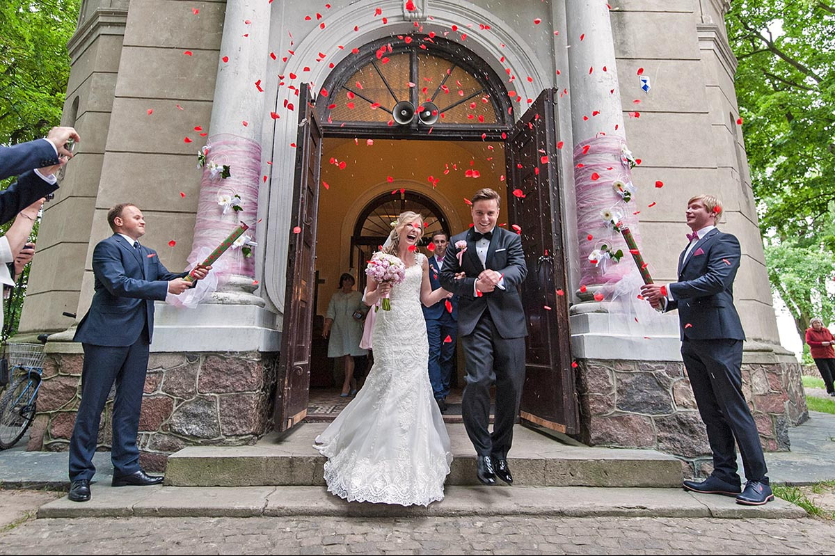 Uśmiechnięta para młoda wychodzi z kościoła, obsypywana płatkami róż. Mężczyźni w garniturach odpalają tuby z konfetti. Dekoracja kwiatowa na kolumnach.