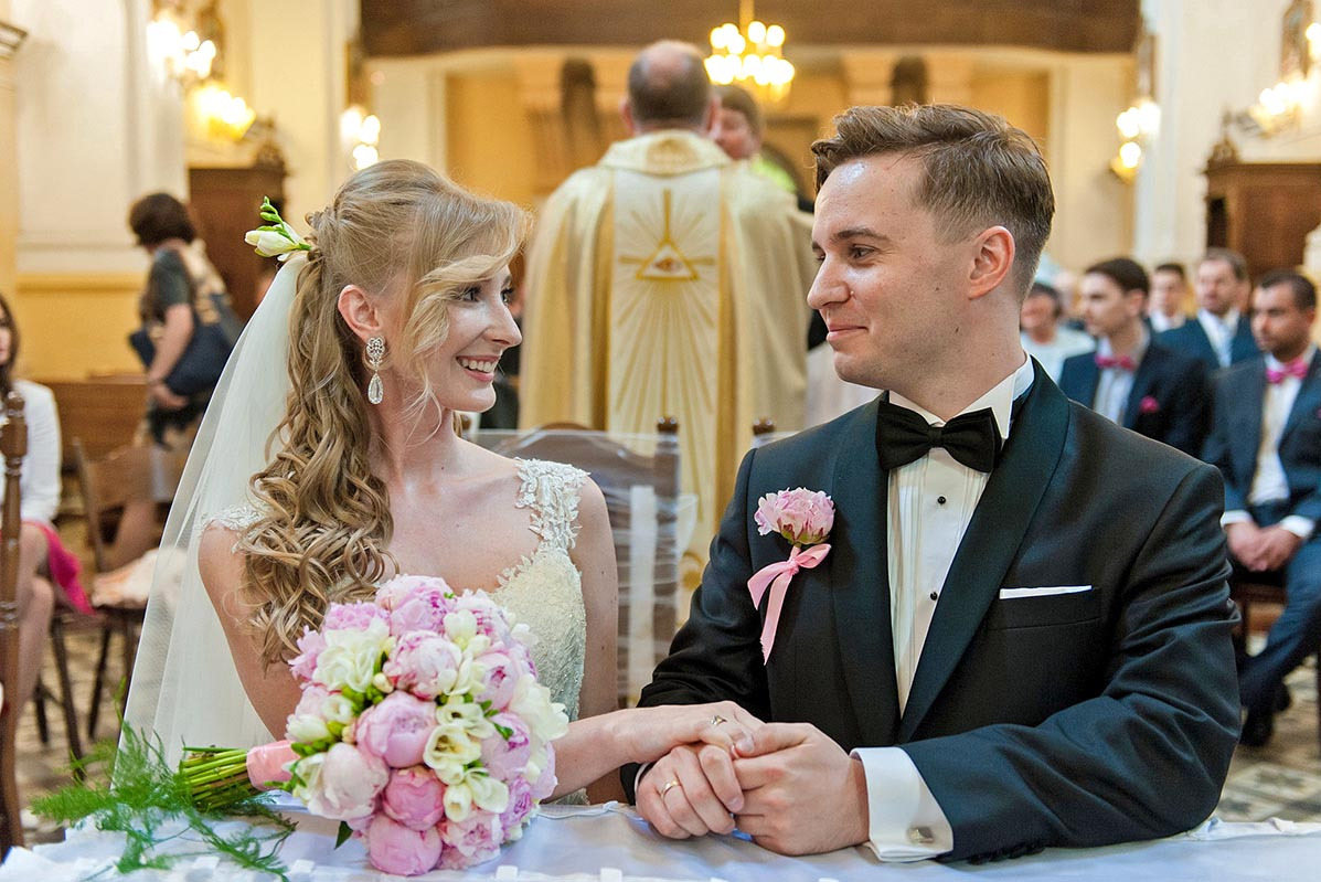 Uśmiechnięta para młoda w kościele, trzymająca się za ręce na tle ołtarza i gości. Bukiet piwonii na pierwszym planie. Elegancki garnitur i suknia ślubna.
