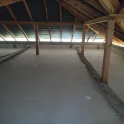 Ocieplone poddasze w trakcie prac. Widoczne drewniane elementy konstrukcji, ułożone płyty izolacyjne na podłodze i fragment ściany z pustaków. Etap prac wykończeniowych.