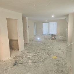 Łukasz Bomba - Wnętrze w trakcie prac wykończeniowych: świeżo pomalowane ściany, zakryta podłoga, widoczny sprzęt budowlany i drabina. Perspektywa z niskiego kąta.