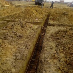 Łukasz Bomba - Zbrojenie wykopu pod fundamenty, widok z góry. W tle maszyny budowlane i osoby. Wykop wąski, zbrojenie z metalowych prętów. Teren budowy, ziemia.