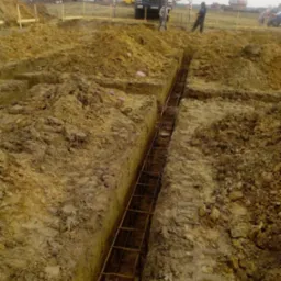 Zbrojenie wykopu pod fundamenty, widok z góry. W tle maszyny budowlane i osoby. Wykop wąski, zbrojenie z metalowych prętów. Teren budowy, ziemia.