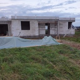 Łukasz Bomba - Surowy stan domu z białego bloczka, widoczne otwory okienne i zbrojenia, przykryty plandeką na tle łąki i pochmurnego nieba. Budowa w toku.
