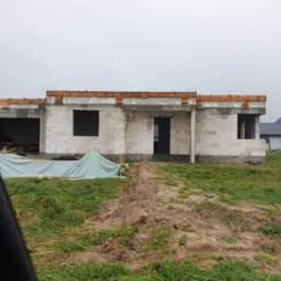 Surowy stan domu z białych bloczków, z drewnianymi elementami stropu i częściowo przykryty plandeką, na tle błotnistego terenu i pochmurnego nieba. Widoczny fragment innego domu w tle.