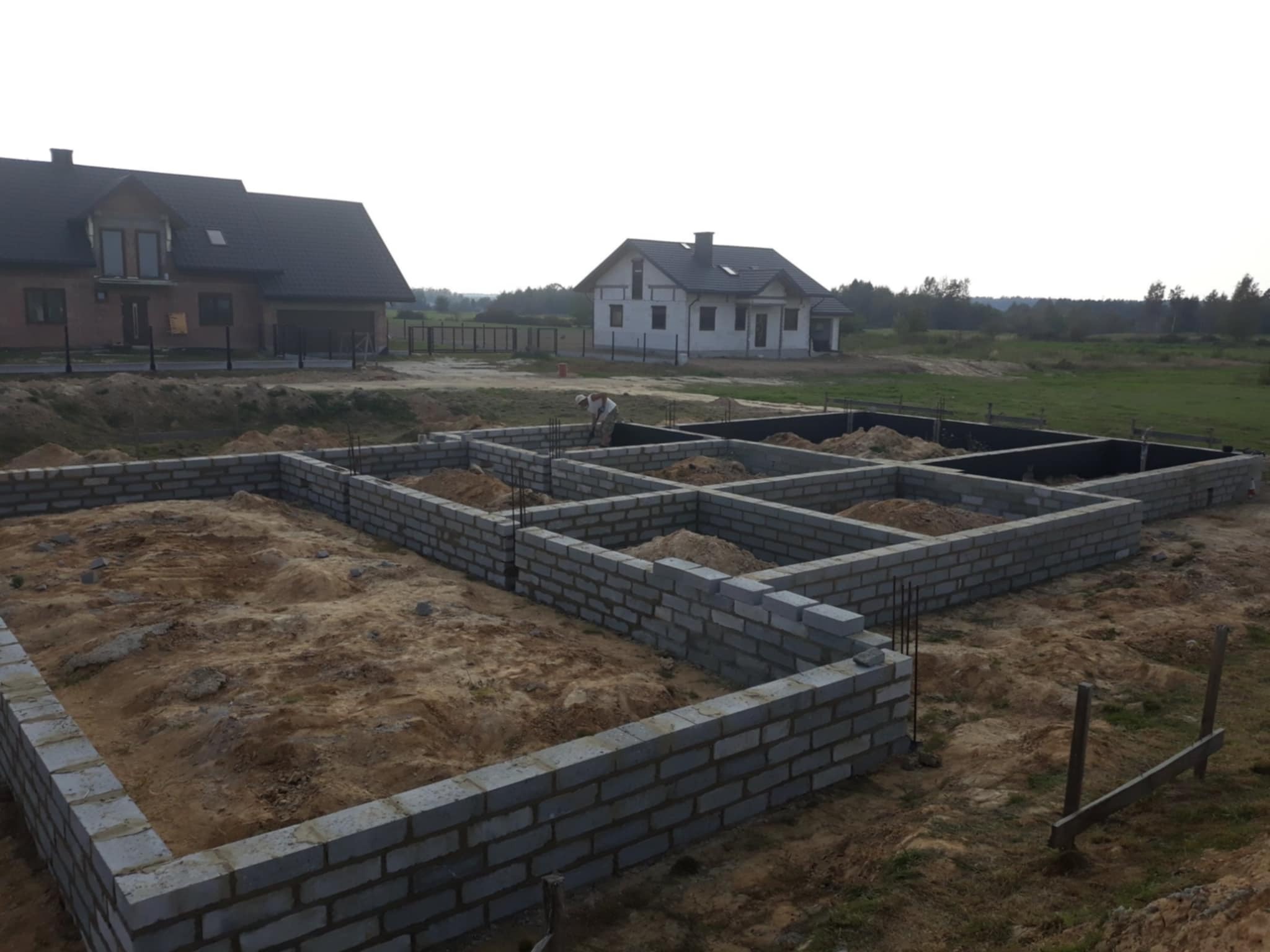 Wykopane i wymurowane fundamenty z bloczków betonowych pod budowę domu. W tle dwa domy jednorodzinne i pracownik budowlany. Widok ogólny.
