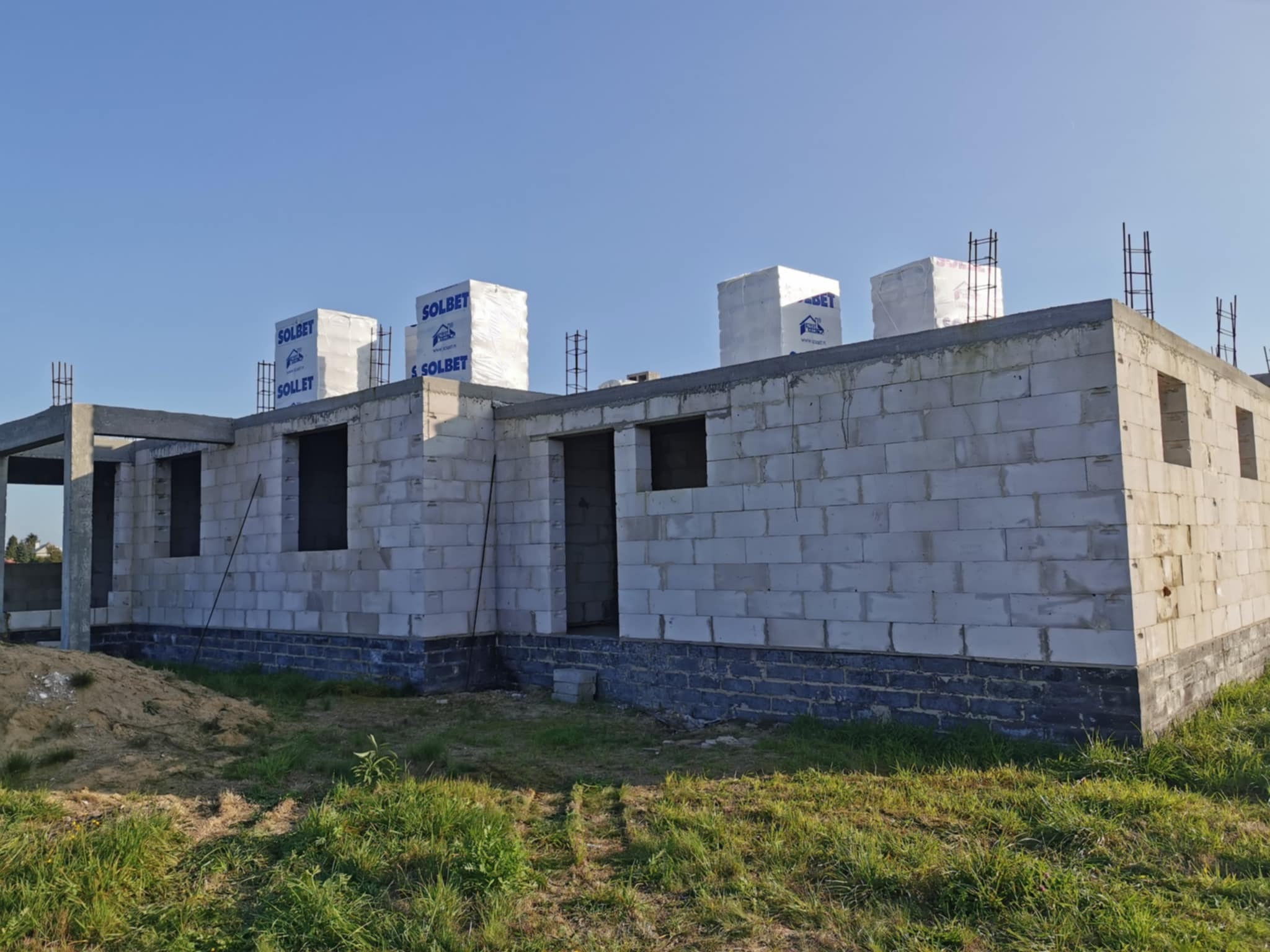 Niedokończony dom z białych bloczków Solbet. Widoczne otwory okienne i drzwiowe, wystające pręty zbrojeniowe. Na bloczkach leżą zapakowane materiały budowlane.