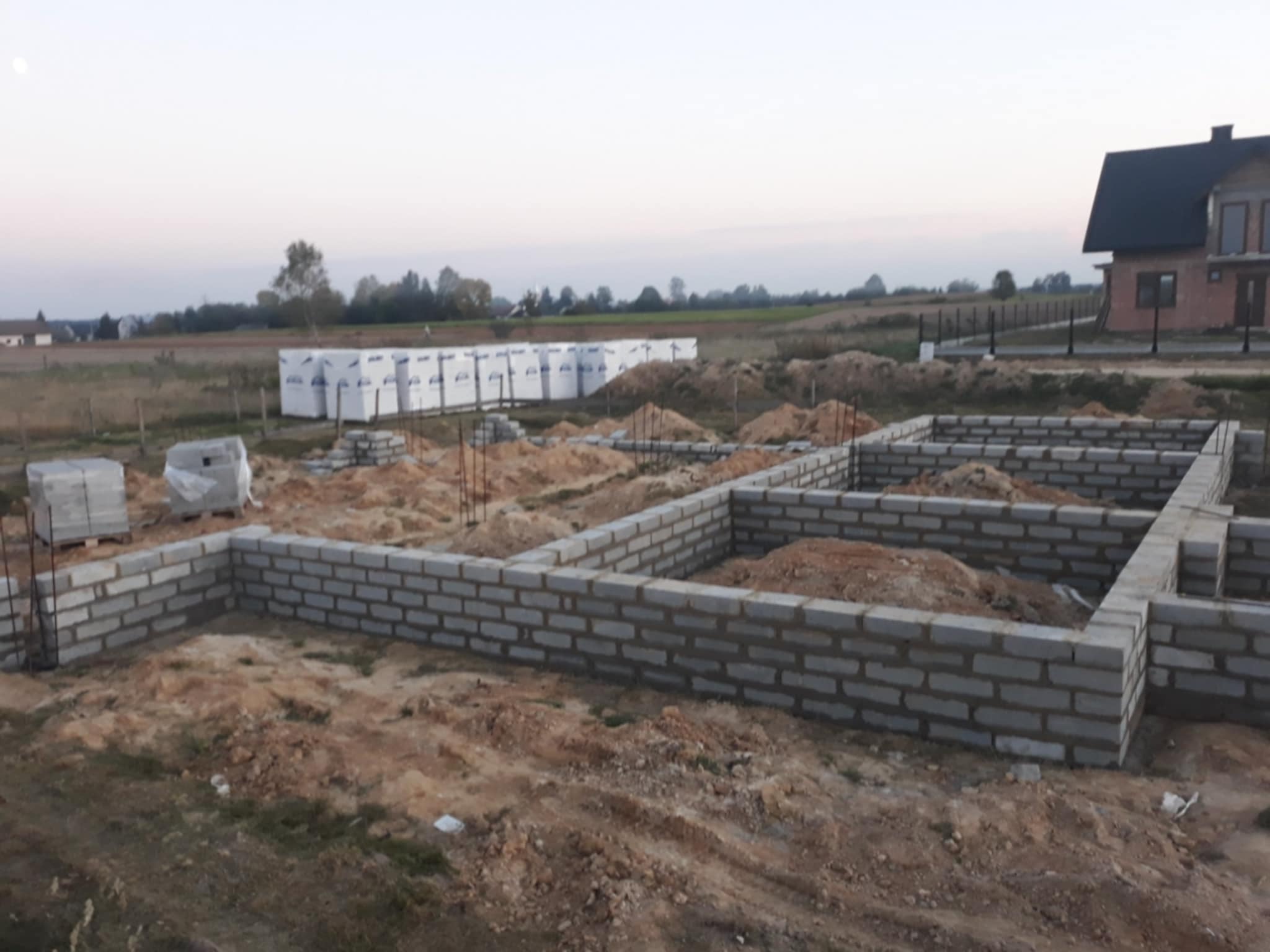 Wzniesione ściany fundamentowe z bloczków betonowych na działce budowlanej. Widoczne zbrojenie i zapasy materiałów. W tle dom w trakcie budowy.