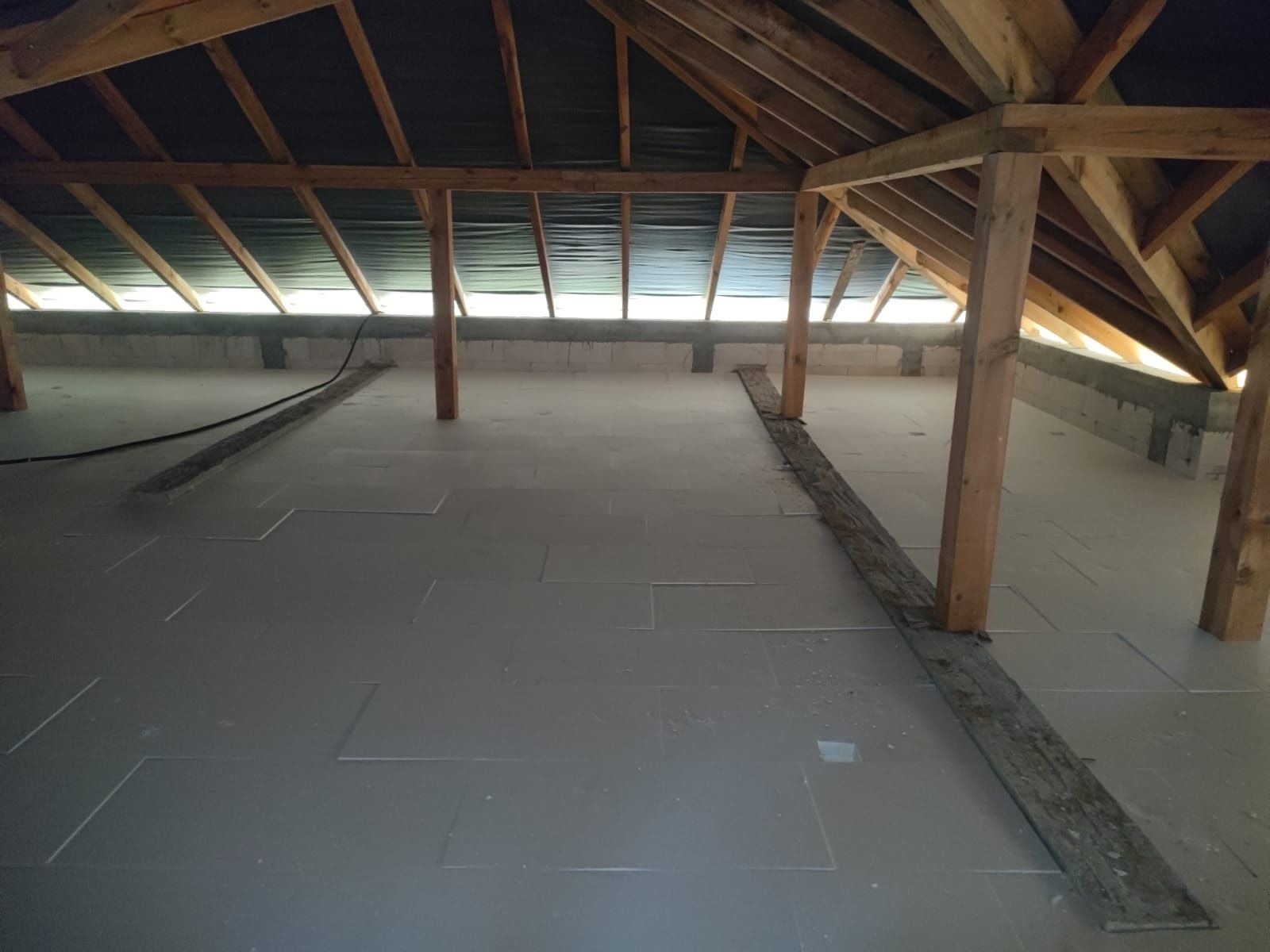 Ocieplone poddasze w trakcie prac. Widoczne drewniane elementy konstrukcji, ułożone płyty izolacyjne na podłodze i fragment ściany z pustaków. Etap prac wykończeniowych.