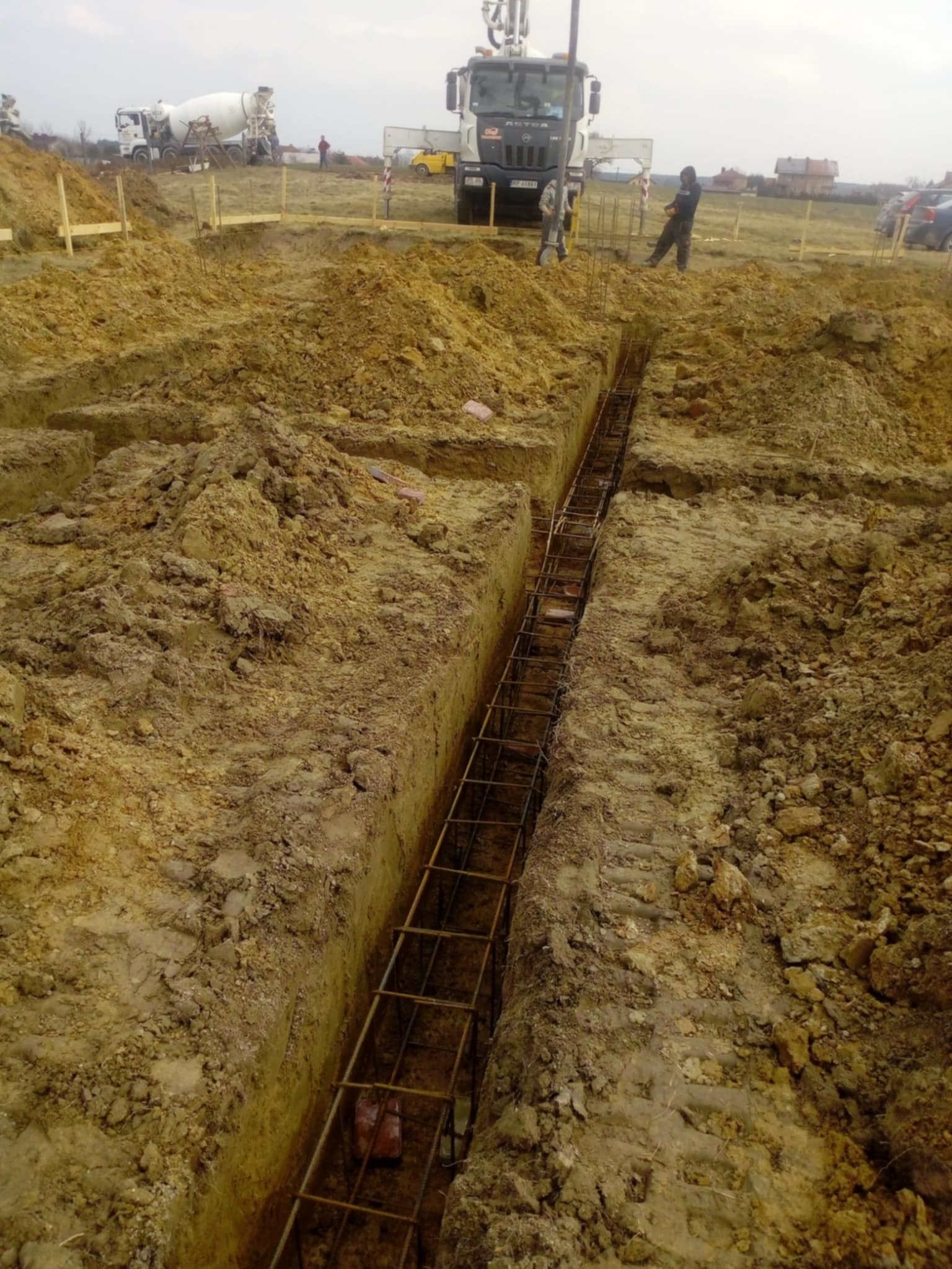 Zbrojenie wykopu pod fundamenty, widok z góry. W tle maszyny budowlane i osoby. Wykop wąski, zbrojenie z metalowych prętów. Teren budowy, ziemia.