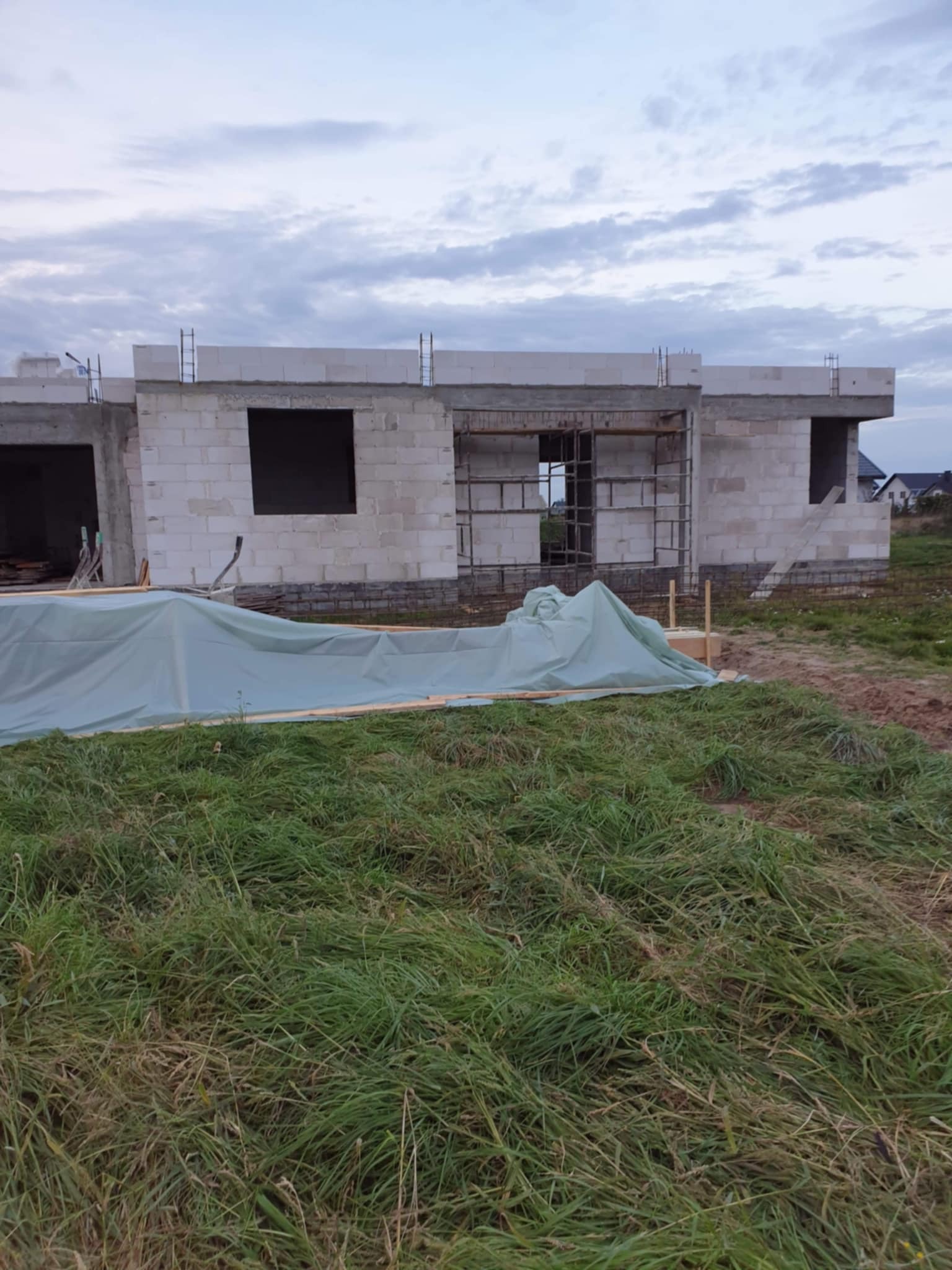 Surowy stan domu z białego bloczka, widoczne otwory okienne i zbrojenia, przykryty plandeką na tle łąki i pochmurnego nieba. Budowa w toku.