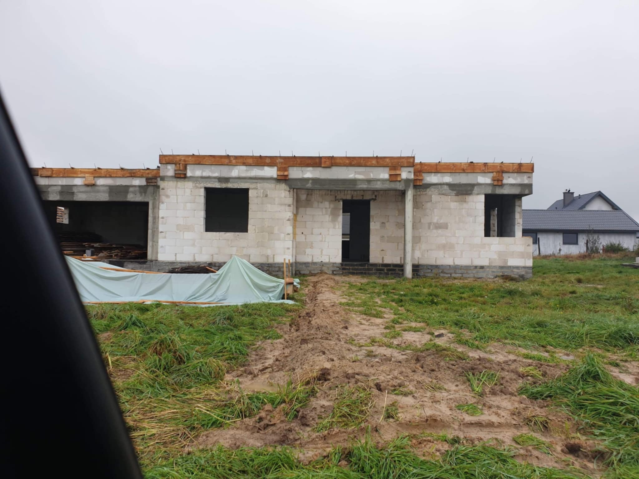 Surowy stan domu z białych bloczków, z drewnianymi elementami stropu i częściowo przykryty plandeką, na tle błotnistego terenu i pochmurnego nieba. Widoczny fragment innego domu w tle.