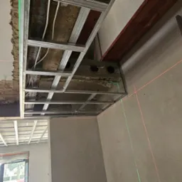 Montaż metalowej konstrukcji sufitu podwieszanego z widocznymi przewodami, poziomica laserowa i drabina oparta o ścianę. W tle okno.